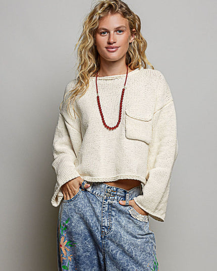 A woman modeling a cream-colored, cropped, long-sleeve knit sweater with a chest pocket, paired with high-waisted blue jeans and a red beaded necklace, against a solid gray studio background.