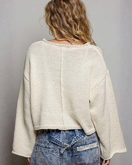 A woman viewed from behind wearing a textured cream knit sweater and blue jeans against a plain gray background.