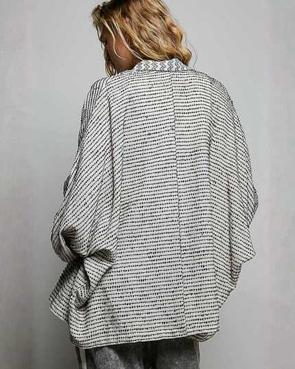 A woman viewed from behind wearing a loose, oversized black and white patterned shirt with wide draped sleeves, paired with gray pants, against a neutral gray wall.
