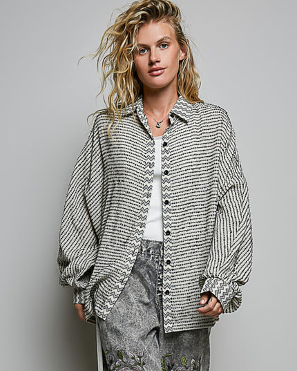 A woman modeling a black and white patterned button-up shirt with balloon sleeves, worn open over a white top and paired with gray patterned jeans, against a solid gray studio background.