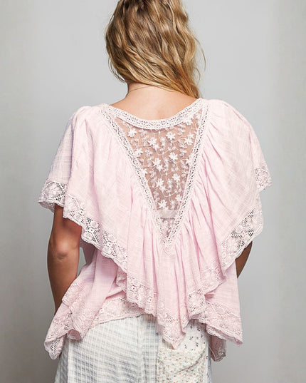 A woman viewed from the back wearing a light pink gauze top with a sheer floral lace yoke and layered ruffled edges, paired with white patterned bottoms, against a plain light gray studio background.