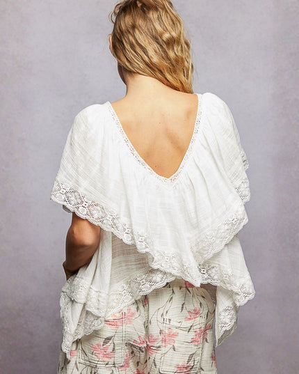 A woman shown from behind wearing a white gauze top with a deep V-back, layered ruffle edges, and lace trim, paired with floral pants against a textured gray studio background.