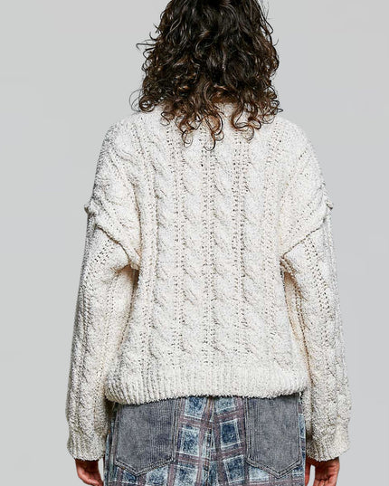 A person shown from the back wearing an oversized cream cable-knit sweater and patterned denim jeans against a plain light gray studio background.