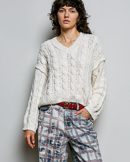 A model wearing a chunky cable-knit cream sweater, paired with a red patterned belt and blue plaid trousers, standing against a light gray studio background.