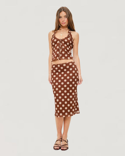 A woman modeling a two-piece brown and beige polka dot set consisting of a halter crop top and a matching midi skirt, paired with brown strappy sandals against a light gray background.