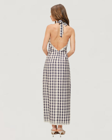 A woman viewed from the back wearing a sleeveless halter-neck maxi dress with a black and white gingham check pattern and an open back, paired with beige high-heeled sandals against a light gray studio background.