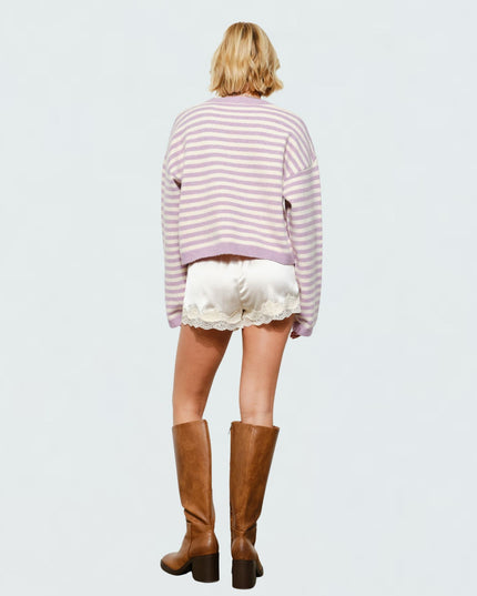 A model viewed from behind wearing a lavender and white striped cropped sweater, white satin shorts with lace trim, and brown knee-high leather boots, set against a plain light gray background.