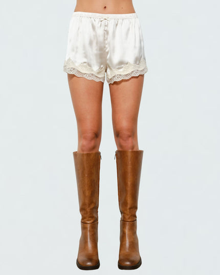 A model's lower body wearing cream satin shorts with a lace hem and brown knee-high leather boots, shown against a light gray studio background.