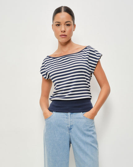 A woman modeling a navy and white horizontally striped off-the-shoulder top with ruffled cap sleeves, paired with light blue jeans, against a plain light gray studio background.