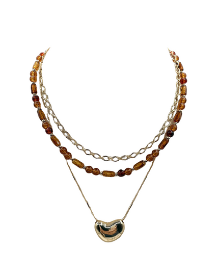 A layered necklace featuring a gold-tone chain with translucent amber beads, a gold-tone chain with clear faceted beads, and a gold-tone chain with a polished gold heart-shaped pendant, set against a plain white background.