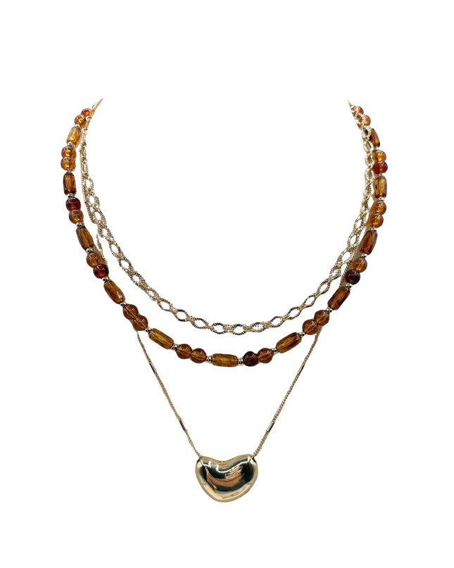 A layered necklace featuring a gold-tone chain with translucent amber beads, a gold-tone chain with clear faceted beads, and a gold-tone chain with a polished gold heart-shaped pendant, set against a plain white background.
