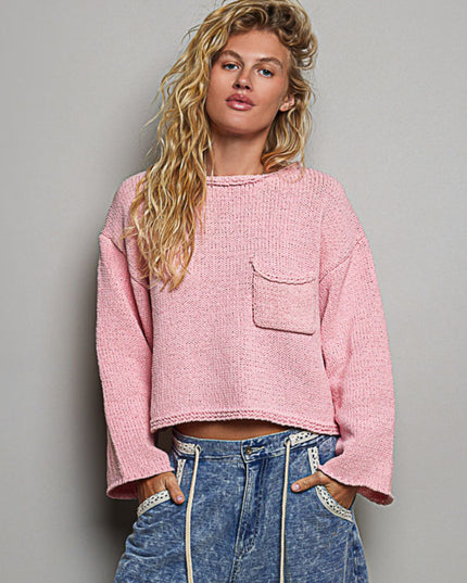 A woman modeling a cropped, textured pink knit sweater with a chest pocket, paired with high-waisted blue jeans, against a solid gray studio background.