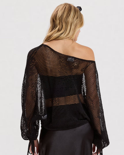 A woman shown from the back wearing a black off-the-shoulder mesh shrug or poncho over a black top and dark skirt, against a plain light gray studio background.