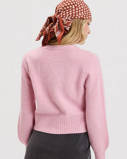 A woman shown from the back wearing a light pink knit sweater, a rust-colored patterned headscarf, and dark gray pants against a plain light gray studio background.