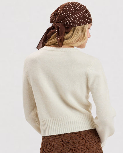 A woman shown from the back wearing a cream knit sweater, a brown polka-dot headscarf, and a brown textured skirt against a plain light gray studio background.