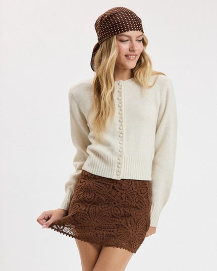 A woman modeling a cream knit sweater with decorative buttons down the front, paired with a brown textured mini skirt and a brown polka-dot bucket hat, against a plain white studio background.