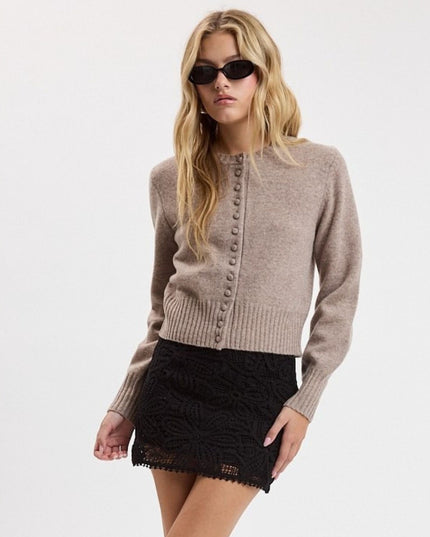 A woman modeling a light taupe knit cardigan sweater with a button-down front and a black lace mini skirt, wearing black sunglasses, against a plain white studio background.
