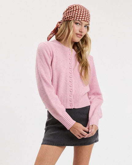 A woman modeling a light pink cropped button-up cardigan sweater, paired with a brown and white checkered headscarf and a black mini skirt, against a plain white studio background.
