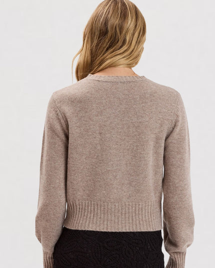 A woman viewed from the back wearing a light taupe knit sweater and a black textured skirt against a plain white studio background.