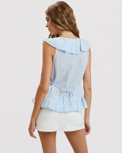 A woman shown from the back wearing a light blue sleeveless top with a ruffled collar and tie detail at the waist, paired with white shorts, against a plain light gray studio background.