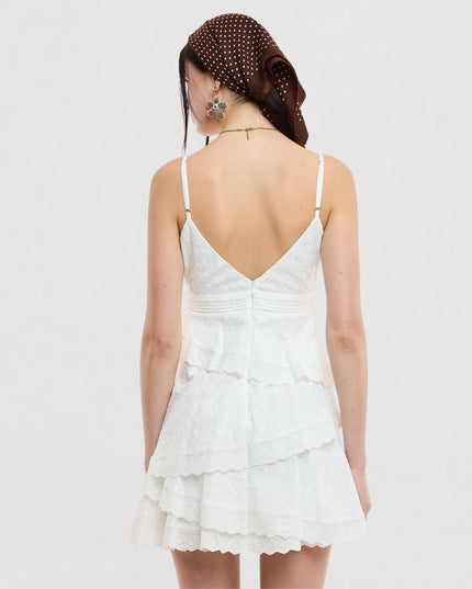 A woman shown from the back wearing a white lace spaghetti-strap mini dress with a ruffled tiered skirt and a brown polka-dot headscarf, against a plain light gray background.