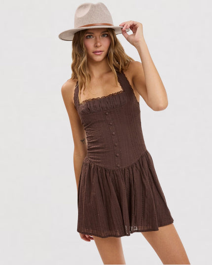 A woman wearing a brown sleeveless mini dress with a ruffled bodice and button front, paired with a beige wide-brim hat, photographed against a light gray studio background.