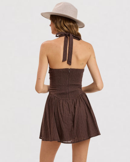 A woman shown from the back wearing a brown halter mini dress with a smocked bodice and a pleated flared skirt, paired with a light beige wide-brim hat against a plain white background.