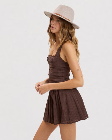 A woman wearing a brown sleeveless mini dress with a pleated skirt and a light beige wide-brim hat, photographed against a plain light gray studio background.
