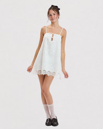 A young woman modeling a white sleeveless mini dress with thin straps, a keyhole cutout at the chest, and a scalloped lace hem, styled with white socks and black loafers against a plain white studio background.