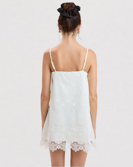 A woman viewed from behind wearing a white spaghetti-strap camisole dress with a lace hem, set against a plain light gray studio background.