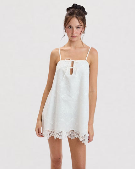 A woman modeling a white spaghetti-strap mini dress with a lace keyhole detail and lace hem, set against a plain light gray studio background.