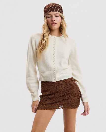 A model wearing a cream ribbed knit cardigan with decorative buttons, a brown textured mini skirt, and a brown patterned beanie against a plain white studio background.