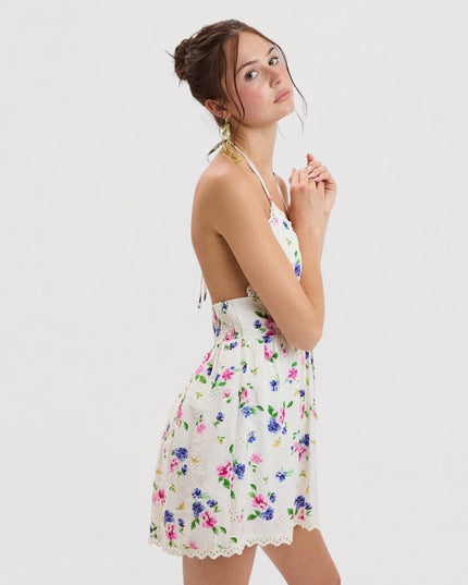 A woman modeling a white halter mini dress with a colorful floral print, set against a plain light gray studio background.
