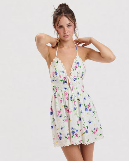 A woman modeling a short white halter mini dress with a colorful floral print and lace trim, set against a plain light gray studio background.