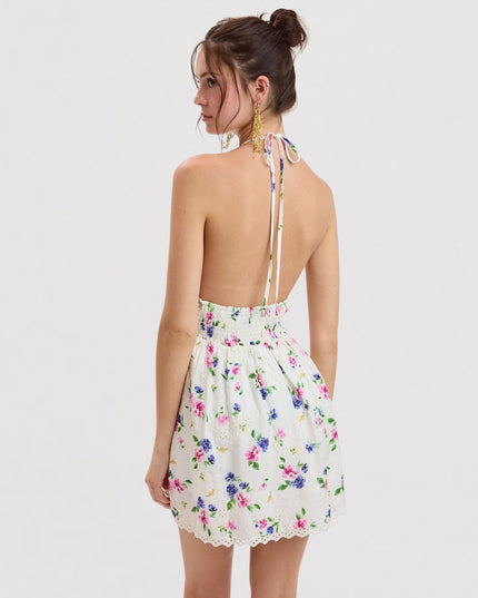 A woman shown from the back wearing a white halter mini dress with a colorful floral print and tie-back straps, photographed against a plain light gray studio background.