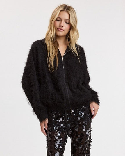 A woman wearing a black fuzzy zip-up jacket and black sequined pants, standing against a plain white studio background.