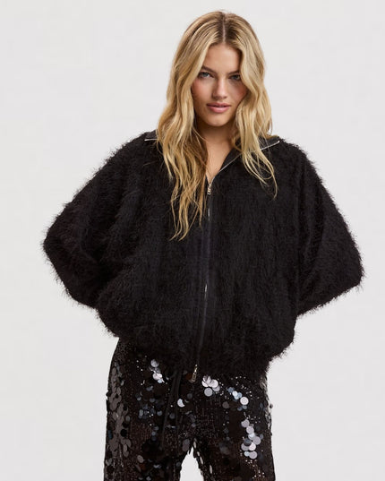 A blonde model wearing a black fuzzy cropped jacket with wide short sleeves and black sequined pants, photographed against a light gray studio background.