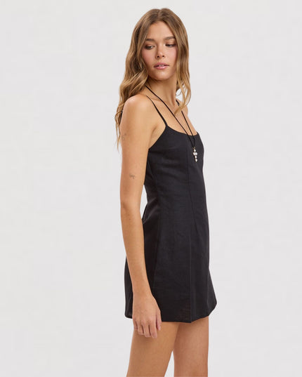 A woman modeling a black spaghetti-strap mini dress against a plain light gray studio background.