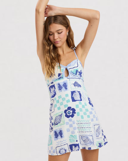 A woman modeling a sleeveless mini dress with a blue and green patchwork print on a white background, featuring spaghetti straps, a keyhole cutout at the chest, and a flared skirt.