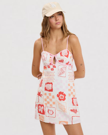 A woman modeling a white spaghetti-strap mini dress with a red and orange tropical patchwork print, paired with a beige baseball cap, against a plain white studio background.