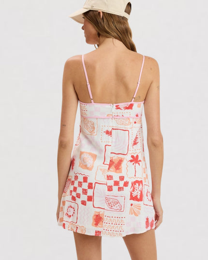 A woman shown from the back wearing a white spaghetti-strap mini dress with a red and orange tropical patchwork print, paired with a beige baseball cap, against a plain white background.