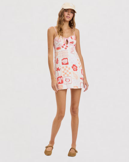 A woman modeling a sleeveless mini dress with a white base and red graphic patchwork print, paired with a beige cap and tan flat sandals, against a light gray studio background.