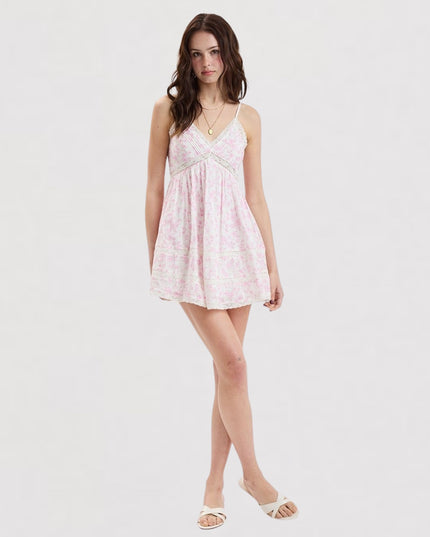 A woman modeling a light pink and white floral print mini dress with thin straps and a gathered skirt, paired with white slide sandals, against a plain white studio background.