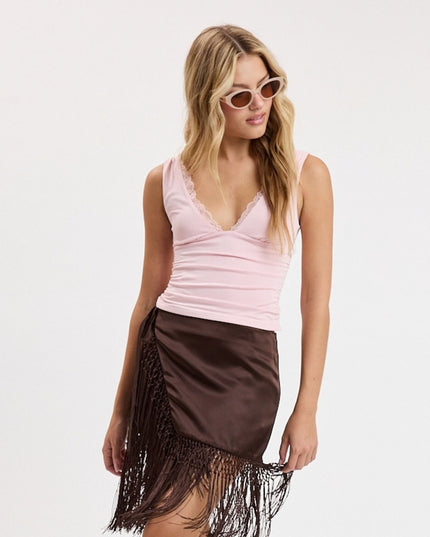 A woman modeling a light pink sleeveless ruched top with a lace-trimmed V-neck and a dark brown satin fringe mini skirt, set against a plain white studio background.
