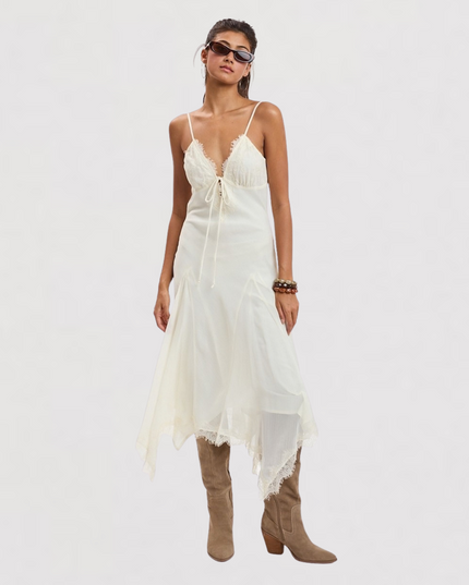 A model wearing a white camisole-style midi dress with lace-up spaghetti straps, a sheer overlay skirt, and frayed hems, styled with brown knee-high boots against a plain white studio background.