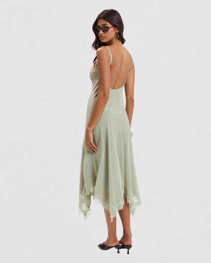 A woman modeling a sage green high-low slip dress with thin straps, a backless open back, and a lace-trimmed hem, shown from the back against a plain white studio background.