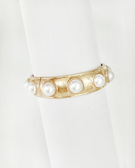 A gold-toned cuff bracelet with five white faux pearls set into a textured channel, displayed on a white cylindrical ring stand.
