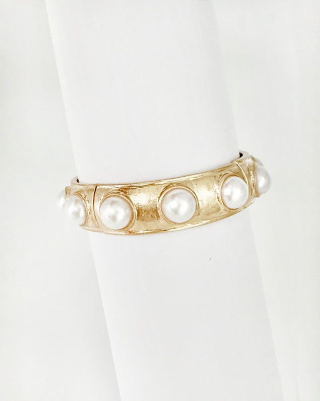 A gold-toned cuff bracelet with five white faux pearls set into a textured channel, displayed on a white cylindrical ring stand.
