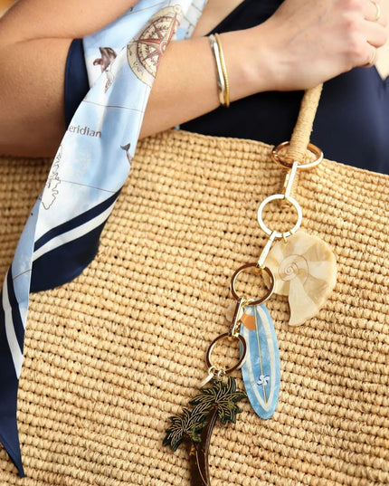 A close-up lifestyle shot of a woven straw tote bag with a decorative keychain strap. The bag features a light blue and navy silk scarf with nautical illustrations, a gold bangle on the wrist, and a keychain with a wooden fan, blue shell, and pinecone charm resting on the bag.