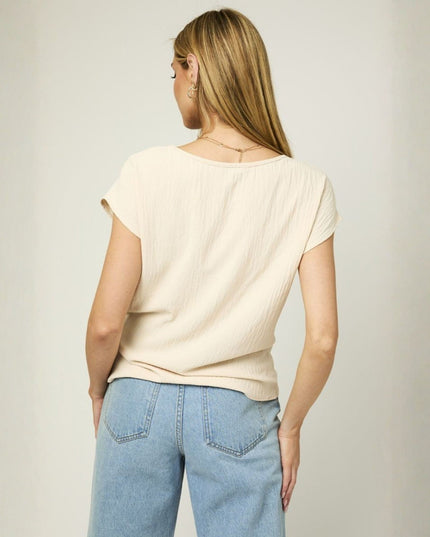 A woman viewed from the back wearing a cream-colored short-sleeve textured top and light blue jeans, standing against a neutral light gray background.